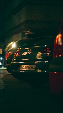 A nighttime scene on a street with several parked cars. A Volkswagen with a visible license plate is at the center, illuminated by streetlights, while other vehicles are partially visible. The surrounding environment is dimly lit, contributing to a moody ambiance.