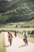 A group of people are cycling along a narrow path amidst a lush, green landscape with terraced fields and dense forested hills in the background. The cyclists are dressed in casual attire and helmets, enjoying the sunny day.