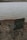 A folding chair sits on a rocky lakeshore. The water is calm, with gentle ripples extending to the distant horizon. The sky is overcast, casting a muted light over the scene.