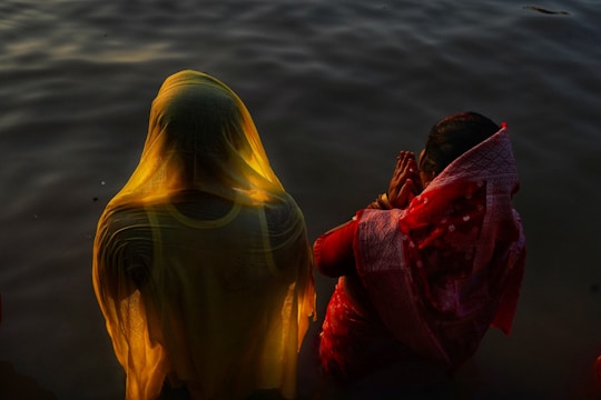 Two individuals are standing at the edge of a body of water. One person is wearing a yellow garment that covers their head and back, creating a soft glow from the sunlight. The other person is dressed in a red and white sari, with hands clasped together in a gesture of prayer or reverence.