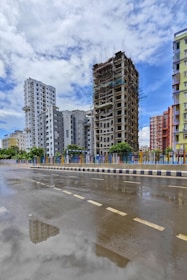 A partially constructed high-rise building surrounded by completed residential buildings under a bright sky with scattered clouds. The street in the foreground reflects the buildings due to recent rainfall, and colorful fencing lines the sidewalk.