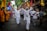 A group of people are participating in a lively street procession, with participants wearing traditional attire and carrying colorful flags. The scene captures a sense of celebration and community engagement as they walk barefoot along the road lined with spectators.