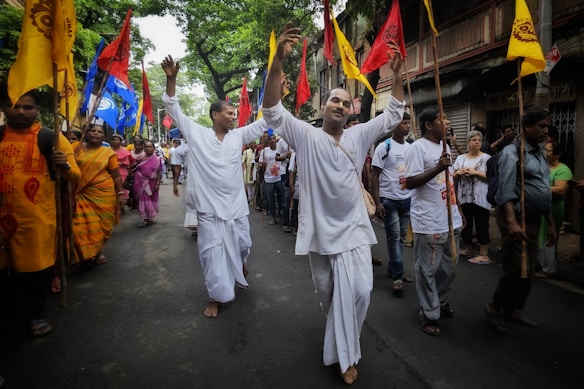 A group of people are participating in a lively street procession, with participants wearing traditional attire and carrying colorful flags. The scene captures a sense of celebration and community engagement as they walk barefoot along the road lined with spectators.