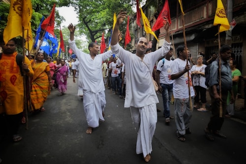 A group of people are participating in a lively street procession, with participants wearing traditional attire and carrying colorful flags. The scene captures a sense of celebration and community engagement as they walk barefoot along the road lined with spectators.