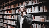 A person in business attire stands in a library, surrounded by shelves filled with books. They appear thoughtful, with one hand touching their neck.