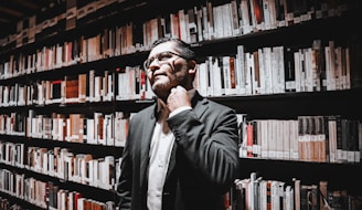 A person in business attire stands in a library, surrounded by shelves filled with books. They appear thoughtful, with one hand touching their neck.