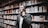 A person in business attire stands in a library, surrounded by shelves filled with books. They appear thoughtful, with one hand touching their neck.