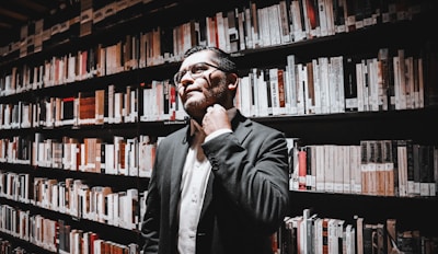 A person in business attire stands in a library, surrounded by shelves filled with books. They appear thoughtful, with one hand touching their neck.