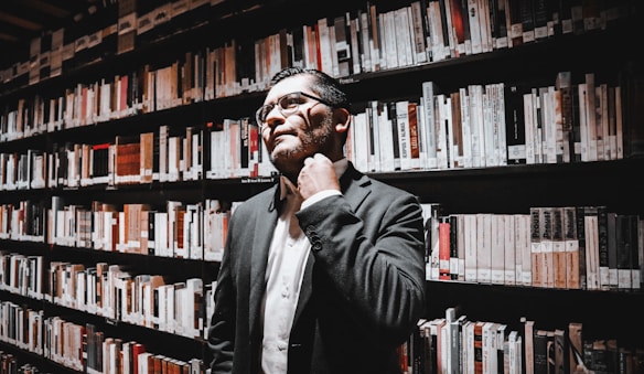 A person in business attire stands in a library, surrounded by shelves filled with books. They appear thoughtful, with one hand touching their neck.