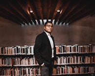 A person wearing a suit stands confidently in a library, surrounded by bookshelves filled with various books. The lighting is warm, and the wooden ceiling beams add depth to the space.
