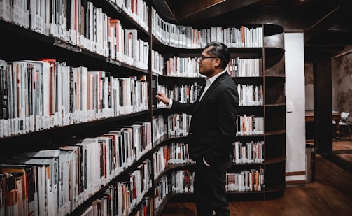 A man in a dark suit is standing in a library, looking at a shelf filled with books. The shelves are curved, extending along the wall, and the man appears to be examining the spines of the books, possibly searching for a specific title. The library has a warm and cozy atmosphere with wooden floors and subtle lighting.