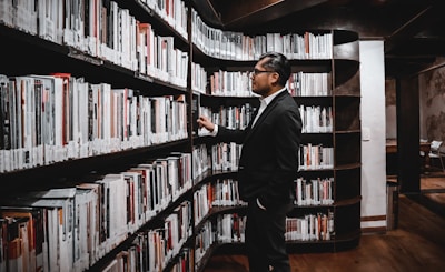 A man in a dark suit is standing in a library, looking at a shelf filled with books. The shelves are curved, extending along the wall, and the man appears to be examining the spines of the books, possibly searching for a specific title. The library has a warm and cozy atmosphere with wooden floors and subtle lighting.