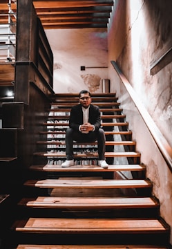 A person wearing a suit sits on a wooden staircase with a wall on one side and bookshelves partially visible behind them. The setting appears warm due to the lighting and textures of the wood and walls.
