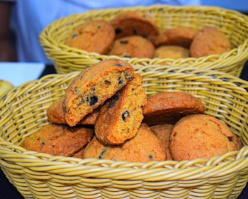 A wicker basket contains a pile of freshly baked cookies with chocolate chips. One cookie is broken in half, revealing a soft interior filled with dark chocolate bits. The cookies have a golden-brown color and appear slightly crispy on the edges.
