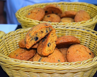 A wicker basket contains a pile of freshly baked cookies with chocolate chips. One cookie is broken in half, revealing a soft interior filled with dark chocolate bits. The cookies have a golden-brown color and appear slightly crispy on the edges.
