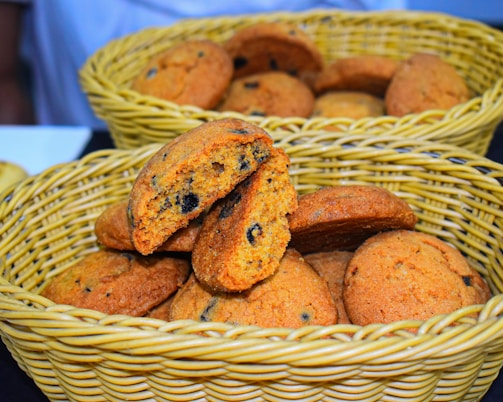 A wicker basket contains a pile of freshly baked cookies with chocolate chips. One cookie is broken in half, revealing a soft interior filled with dark chocolate bits. The cookies have a golden-brown color and appear slightly crispy on the edges.