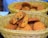 A wicker basket contains a pile of freshly baked cookies with chocolate chips. One cookie is broken in half, revealing a soft interior filled with dark chocolate bits. The cookies have a golden-brown color and appear slightly crispy on the edges.