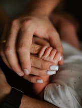 An adult hand gently holding a baby's hand, both resting on a surface. The adult hand has well-manicured nails with white nail polish.