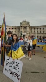 A group of people are gathered in front of a large historical building holding flags and signs. A young woman at the forefront is draped in a blue and yellow flag, holding a sign that reads 'DO NOT SIT AT ONE TABLE WITH MURDERERS'. The atmosphere appears solemn and serious.