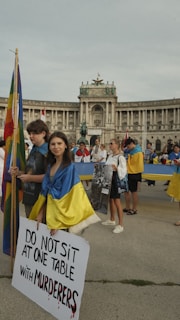 A group of people are gathered in front of a large historical building holding flags and signs. A young woman at the forefront is draped in a blue and yellow flag, holding a sign that reads 'DO NOT SIT AT ONE TABLE WITH MURDERERS'. The atmosphere appears solemn and serious.