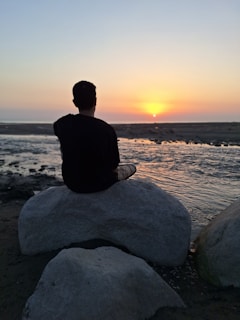 A person meditating on a rock overlooking the calm beach at sunset.