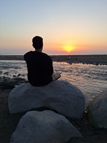 A person meditating on a rock overlooking the calm beach at sunset.