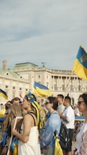 A group of people are gathered outside a large historic building. Many are holding Ukrainian flags, and some are wearing traditional Ukrainian clothing adorned with blue and yellow accents. The building behind features ornate architecture with columns and statues. The sky is clear, suggesting a sunny day.
