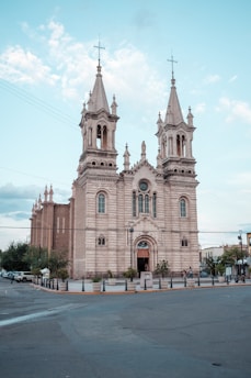 A grand, historic church with two tall towers topped with crosses, featuring intricate architectural details. It is constructed from stone and stands prominently at the intersection of a road, surrounded by trees and a few people walking nearby under a bright blue sky with scattered clouds.