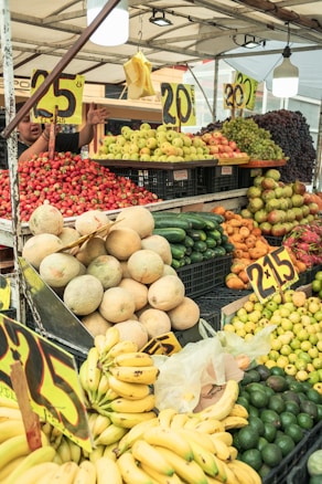 A vibrant market stall displays a variety of colorful fruits, including bananas, avocados, melons, cucumbers, strawberries, apples, and grapes. Price signs indicating the cost per kilogram are prominently featured. A vendor can be seen attending to the fruit arrangement in the background.
