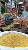 An indoor market stall is filled with a variety of spices and dried goods. There are transparent containers and bags holding items like walnuts, green herbs, lentils, nuts, and various spices. A person wearing a light green shirt is partially visible, possibly attending to the stall. Shelves in the background are stocked with more packages and jars, indicating a busy market ambiance.