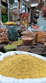 An indoor market stall is filled with a variety of spices and dried goods. There are transparent containers and bags holding items like walnuts, green herbs, lentils, nuts, and various spices. A person wearing a light green shirt is partially visible, possibly attending to the stall. Shelves in the background are stocked with more packages and jars, indicating a busy market ambiance.