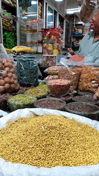 An indoor market stall is filled with a variety of spices and dried goods. There are transparent containers and bags holding items like walnuts, green herbs, lentils, nuts, and various spices. A person wearing a light green shirt is partially visible, possibly attending to the stall. Shelves in the background are stocked with more packages and jars, indicating a busy market ambiance.