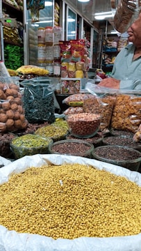 An indoor market stall is filled with a variety of spices and dried goods. There are transparent containers and bags holding items like walnuts, green herbs, lentils, nuts, and various spices. A person wearing a light green shirt is partially visible, possibly attending to the stall. Shelves in the background are stocked with more packages and jars, indicating a busy market ambiance.