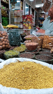 An indoor market stall is filled with a variety of spices and dried goods. There are transparent containers and bags holding items like walnuts, green herbs, lentils, nuts, and various spices. A person wearing a light green shirt is partially visible, possibly attending to the stall. Shelves in the background are stocked with more packages and jars, indicating a busy market ambiance.