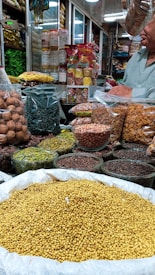 An indoor market stall is filled with a variety of spices and dried goods. There are transparent containers and bags holding items like walnuts, green herbs, lentils, nuts, and various spices. A person wearing a light green shirt is partially visible, possibly attending to the stall. Shelves in the background are stocked with more packages and jars, indicating a busy market ambiance.
