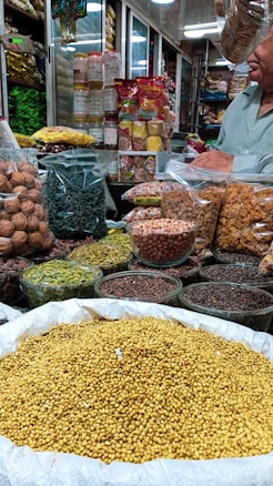 An indoor market stall is filled with a variety of spices and dried goods. There are transparent containers and bags holding items like walnuts, green herbs, lentils, nuts, and various spices. A person wearing a light green shirt is partially visible, possibly attending to the stall. Shelves in the background are stocked with more packages and jars, indicating a busy market ambiance.