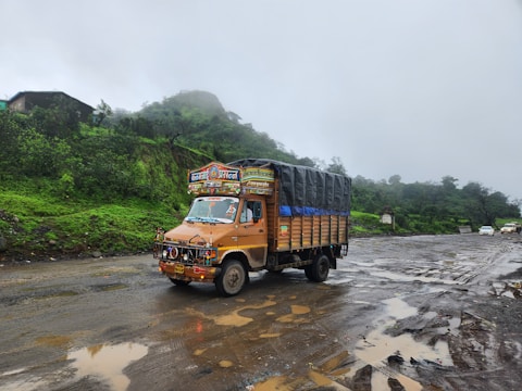 A decorated truck drives along a wet and muddy road. The truck is adorned with colorful patterns and writings on the front. Surrounding the road are lush green hills, partially obscured by mist and fog. A couple of other vehicles are visible in the background.