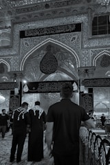 An ornate interior features detailed Arabic calligraphy and decorative arches. People wearing black clothing, some with keffiyehs, walk on a patterned carpet. The scene reflects a solemn and reverent atmosphere.