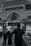 An ornate interior features detailed Arabic calligraphy and decorative arches. People wearing black clothing, some with keffiyehs, walk on a patterned carpet. The scene reflects a solemn and reverent atmosphere.
