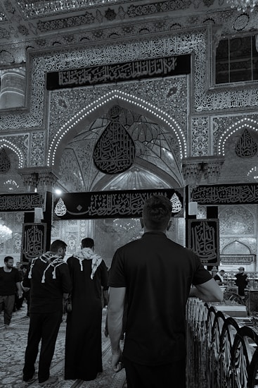An ornate interior features detailed Arabic calligraphy and decorative arches. People wearing black clothing, some with keffiyehs, walk on a patterned carpet. The scene reflects a solemn and reverent atmosphere.