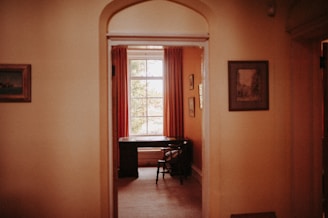 A cozy room viewed through an open doorway, featuring a classic wooden desk and chair near a large window dressed with red curtains. The walls are adorned with framed art, and soft, warm lighting creates an inviting atmosphere.