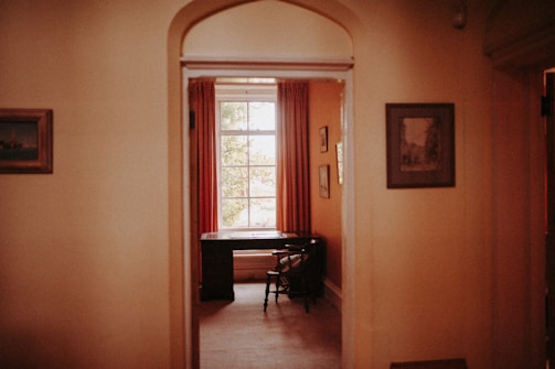 A cozy room viewed through an open doorway, featuring a classic wooden desk and chair near a large window dressed with red curtains. The walls are adorned with framed art, and soft, warm lighting creates an inviting atmosphere.
