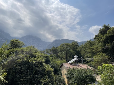 A serene landscape featuring a lush forest with green trees surrounding a small house with solar panels on the roof. In the background, rolling hills and mountains can be seen partially covered by clouds under a partly cloudy sky.