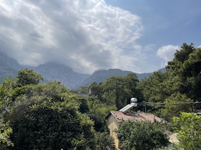 A serene landscape featuring a lush forest with green trees surrounding a small house with solar panels on the roof. In the background, rolling hills and mountains can be seen partially covered by clouds under a partly cloudy sky.