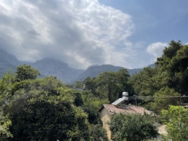 A serene landscape featuring a lush forest with green trees surrounding a small house with solar panels on the roof. In the background, rolling hills and mountains can be seen partially covered by clouds under a partly cloudy sky.