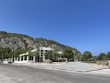 A paved road leads to a residential building surrounded by a white fence. The area is bordered by lush greenery and a rocky hillside. Construction cones and a few cars are visible along the roadside.