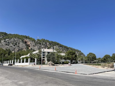 A paved road leads to a residential building surrounded by a white fence. The area is bordered by lush greenery and a rocky hillside. Construction cones and a few cars are visible along the roadside.