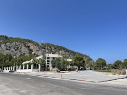 A paved road leads to a residential building surrounded by a white fence. The area is bordered by lush greenery and a rocky hillside. Construction cones and a few cars are visible along the roadside.