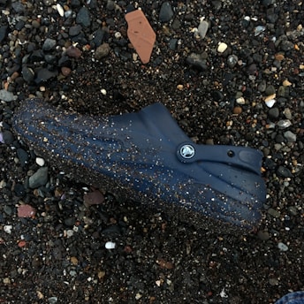 A blue rubber clog is lying on a pebbled beach. The shoe is covered in small particles of sand and surrounded by variously sized stones and rocks. A small piece of terracotta tile is partially buried in the sand nearby.