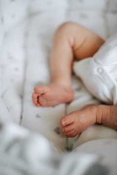 Close-up of a baby's legs and feet resting on a soft, cushioned surface, with a focus on the delicate toes and smooth skin. The background features a light-colored fabric with subtle patterns.